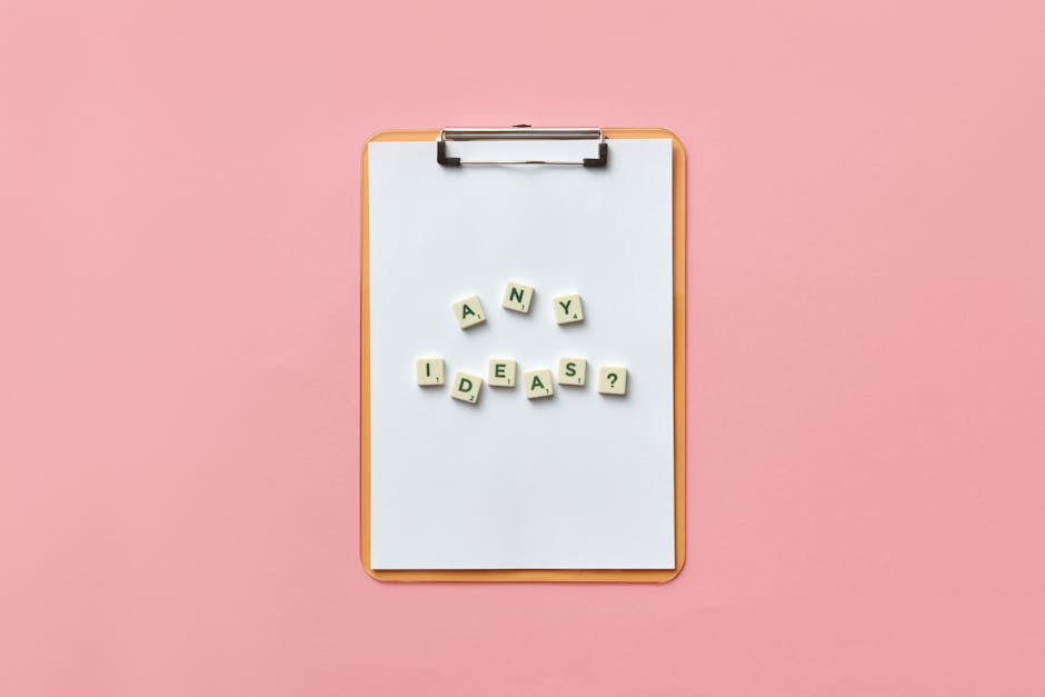 Creative conceptual image of a clipboard with letters spelling 'Any Ideas?' using Scrabble tiles on a pink background.